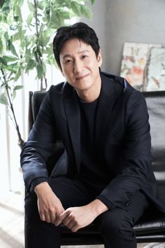 This contains an image of Lee Sun Kyun  sitting on top of a black chair next to a potted green plant