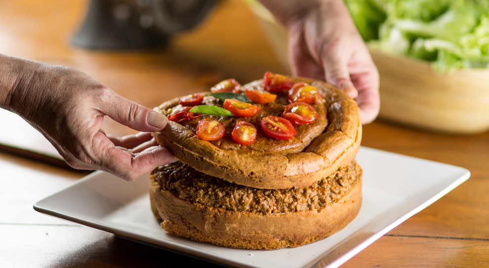 A imagem mostra uma torta mexicana sendo montada. A base é um bolo salgado dourado, recheado com carne moída bem temperada. A parte superior, decorada com molho, tomates-cereja e folhas verdes, está sendo cuidadosamente posicionada sobre o recheio. O prato está em um ambiente rústico, com uma salada ao fundo.