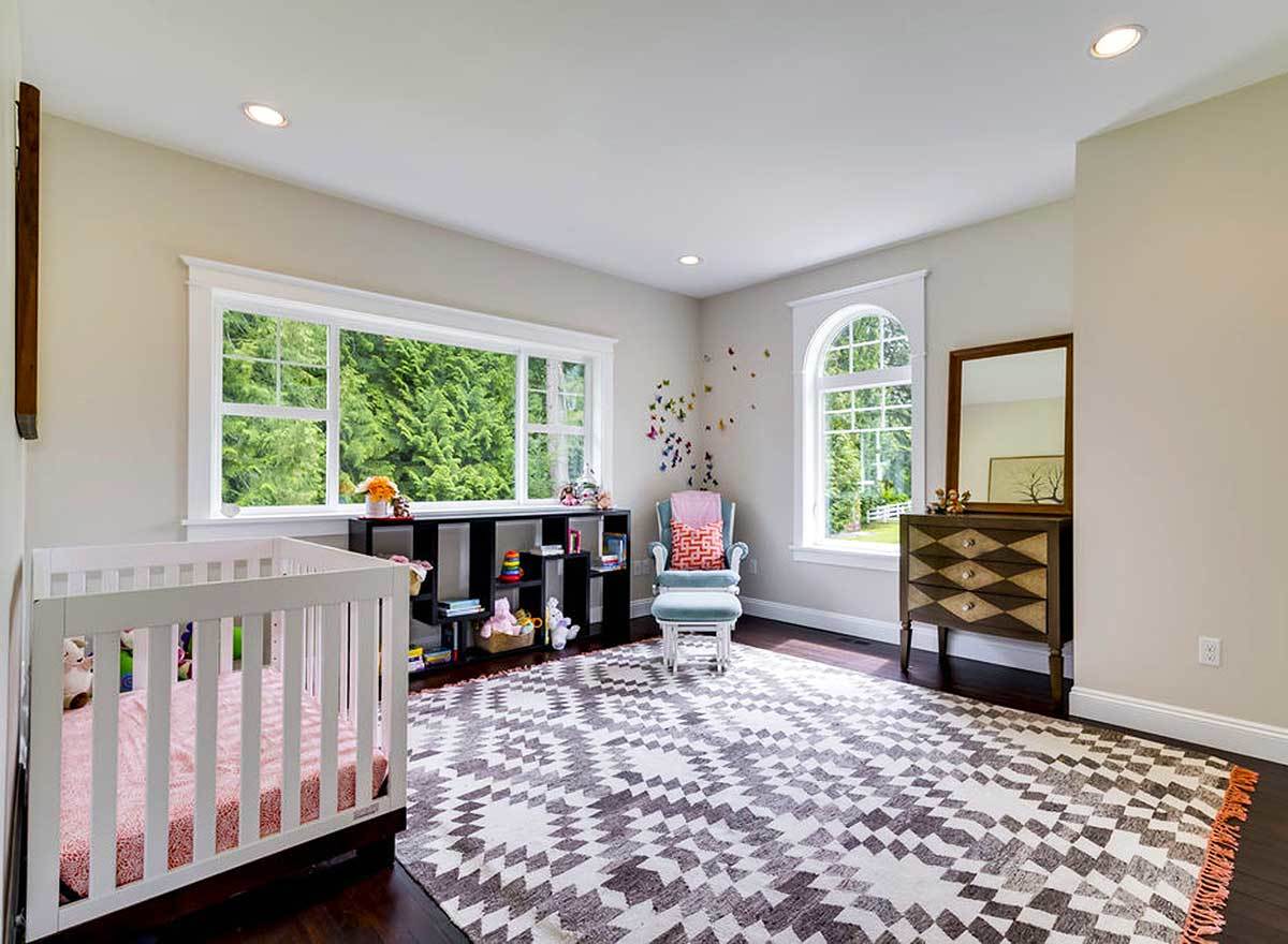 This bedroom has beige walls, a white crib, wooden vanity, and a blue lounge chair over a large patterned rug.
