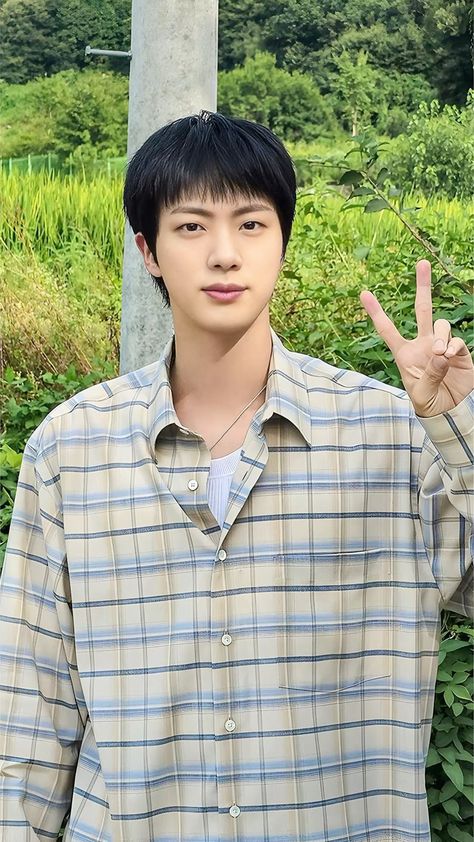 This contains an image of BTS Jin  making the peace sign with his hand while standing in front of some bushes