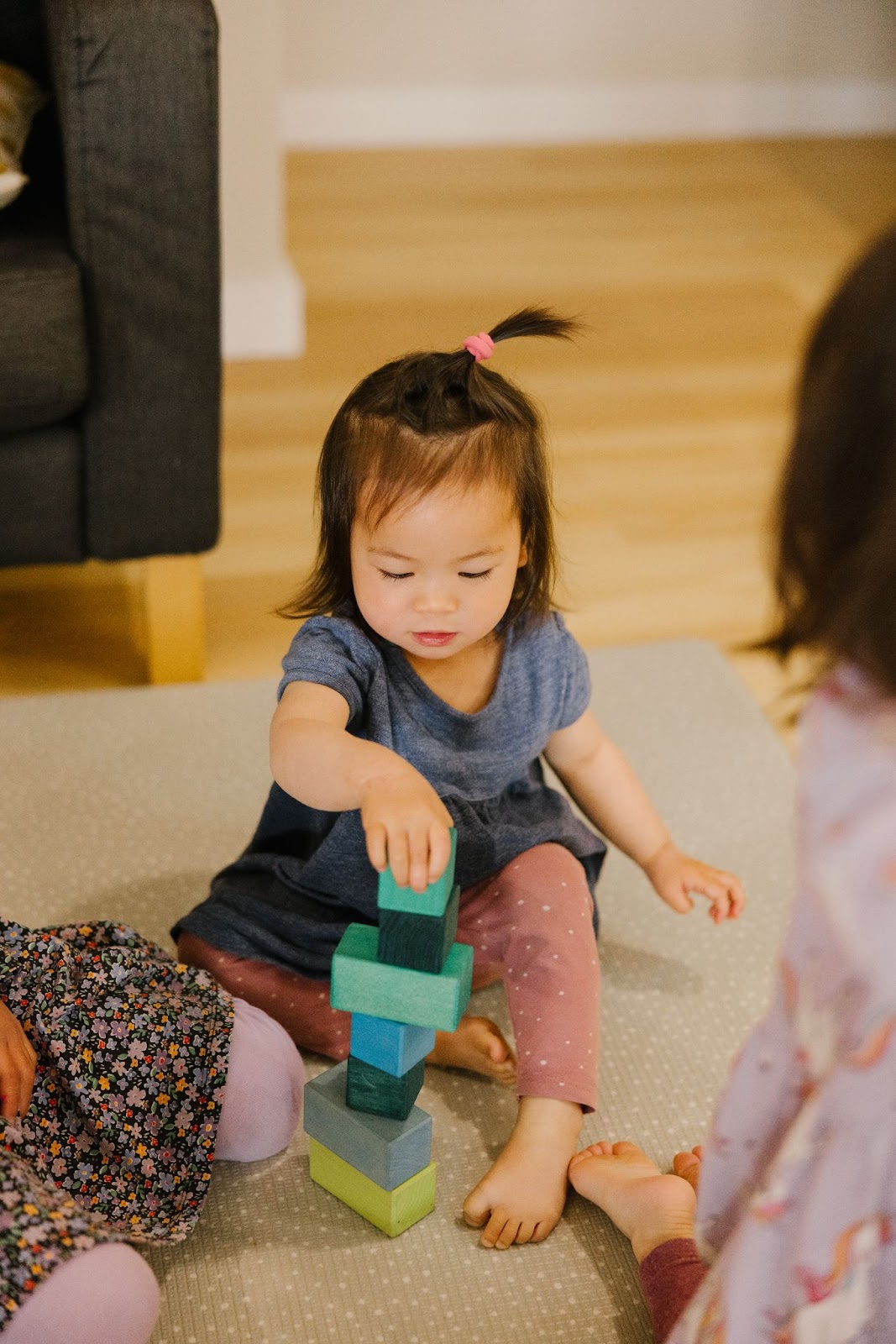 A young toddler carefully picks up a cool-toned block from Grimm’s Large Stepped Pyramid set, and carefully stacks it to build a tower in shades of blue and green.