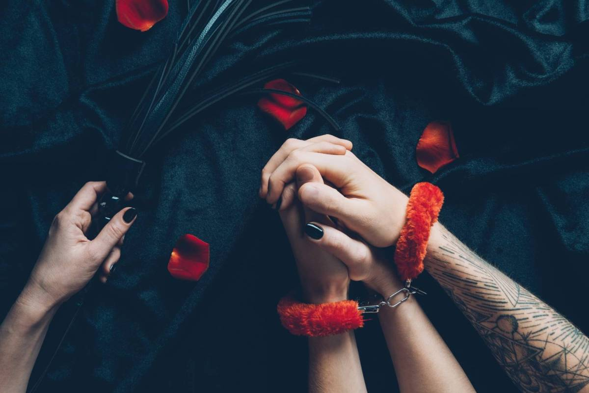 A pair of masculine-looking hands wear red fuzzy handcuffs, while a feminine hand holds a black flogging toy against a black background.