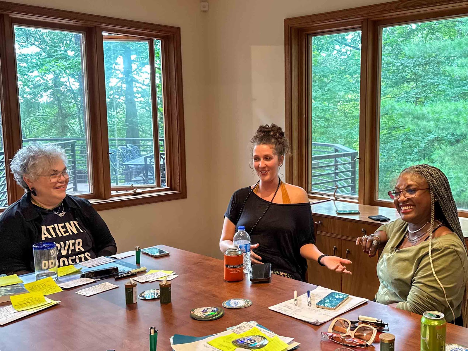 Photo depicts Arkansas Roots & Reefer educational cannabis podcast hosts interviewing grow lead at The Source Cannabis Dispensary and boutique grow, Ari Pascoe, sitting around a wooden table at a home in Northwest Arkansas. Behind the three smiling women are large windows giving us a view of a lush summer forest.