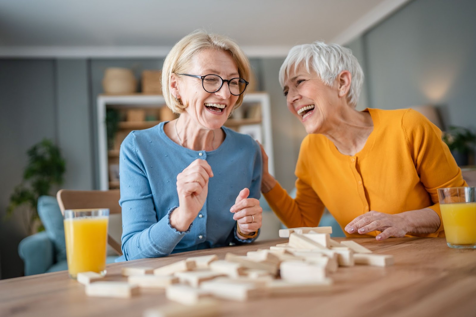 2 older adult women smiling and sitting at a table together playing a game while drinking orange juice.