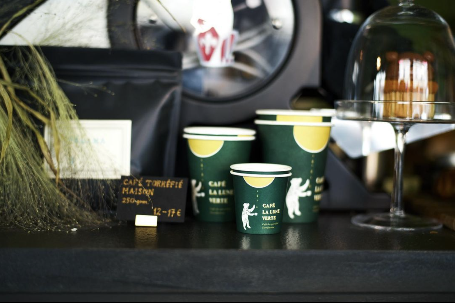 Green branded cups with white bear illustrations displayed on a shelf, surrounded by decorative dried plants, coffee beans, and a glass dome with pastries.