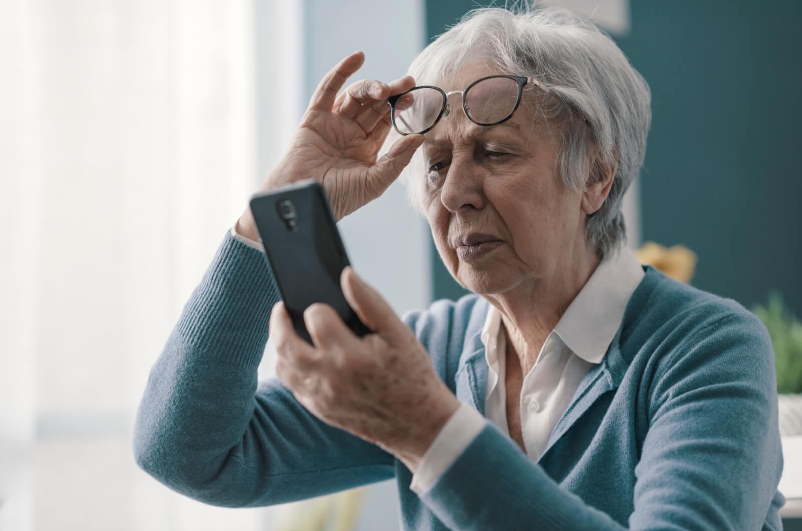 An older adult lifting their outdated prescription glasses above their eyes with their right hand as they squint at their phone held in their left hand