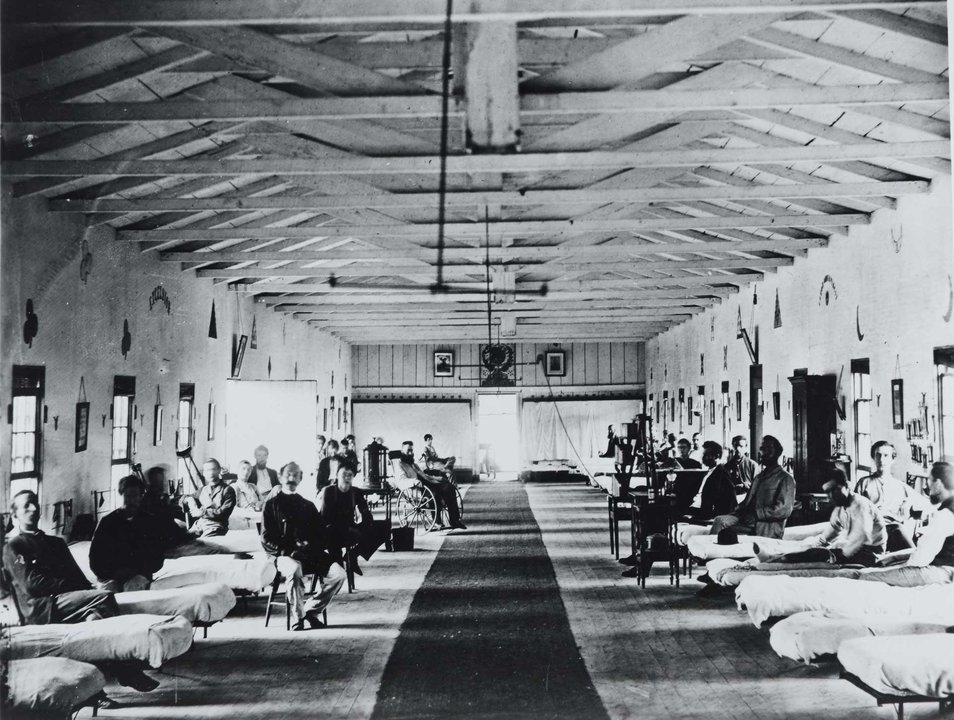 This appears to be a historical hospital ward or medical facility, with a long corridor lined with beds on either side. The room has exposed wooden ceiling beams and a central walkway. Patients and medical staff are visible throughout the space, with some seated in chairs and others resting in beds. The lighting comes from windows along the walls, and there appear to be various medical equipment and furnishings scattered throughout. The image is in black and white, suggesting it was taken in an earlier era, possibly early 20th century.
The architectural style and layout is typical of medical facilities from this time period, with its high ceilings, open ward design, and long rectangular shape that allowed for maximum ventilation and supervision of multiple patients. The walls appear to be decorated with some pictures or artwork, and there's what looks like medical equipment or possibly a wheelchair visible in the background.