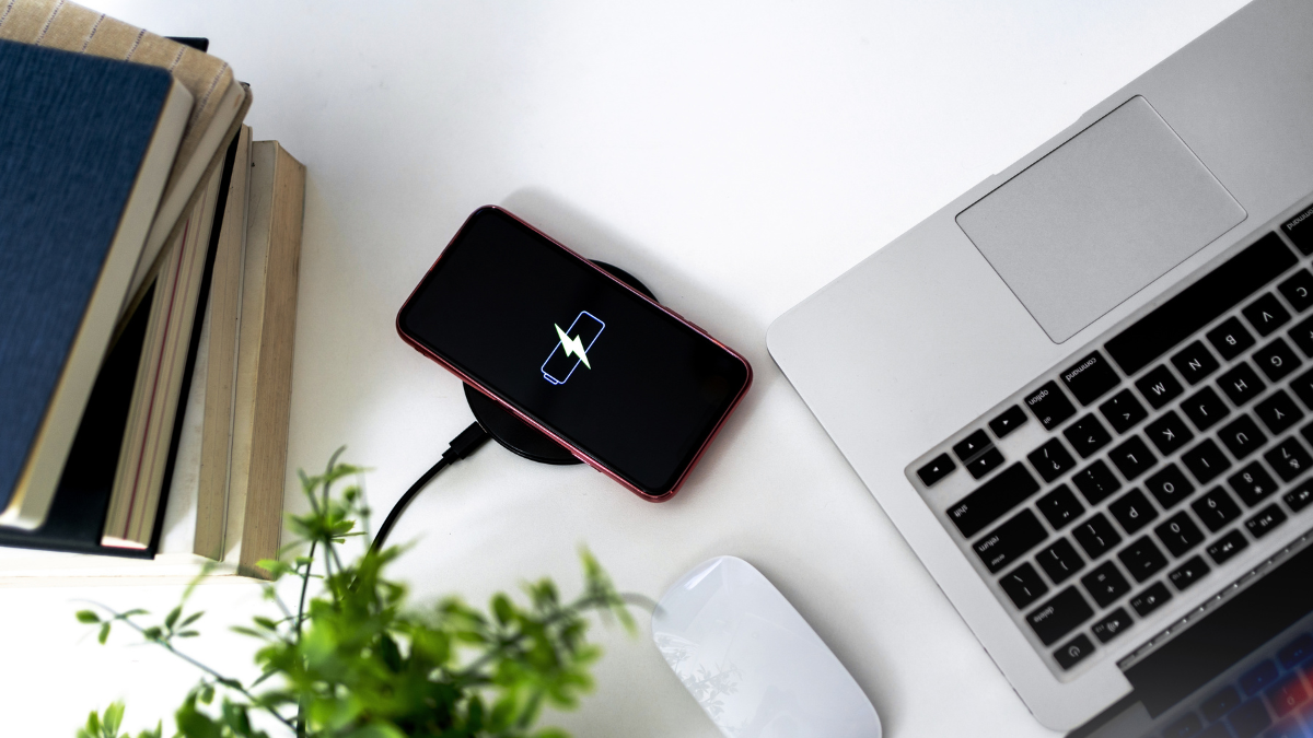 Modern workspace featuring a wireless smartphone charger with a charging phone, a laptop, a mouse, a stack of books, and a green plant on a white desk.