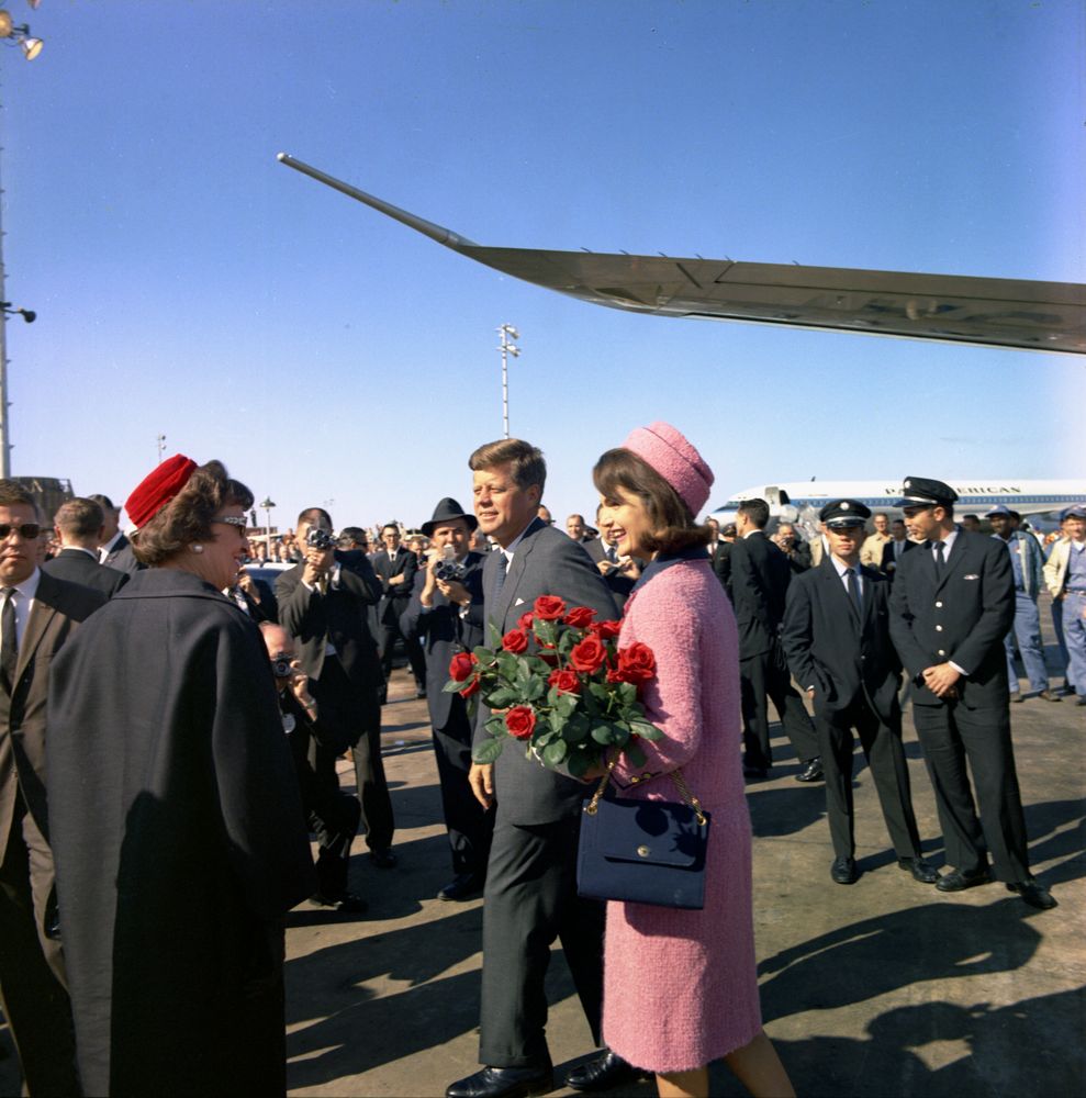 President John F. Kennedy and First Lady Jackie Kennedy greeting citizens at Love Field after touching down in Dallas, Texas. Jackie is seen in her iconic pink Chanel suit, holding a bouquet of red roses.