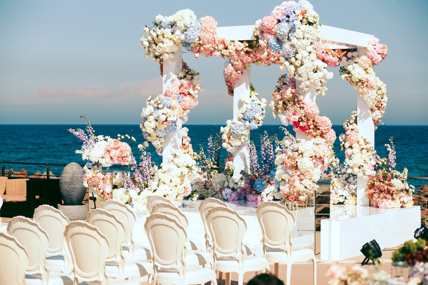 Romantic beach wedding gazebo decorated with flowing white drapes, tropical flowers, and lanterns under a clear blue sky.

