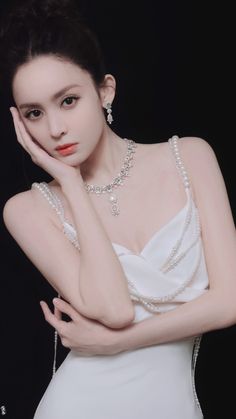 This contains an image of Guli Nazha
white dress with pearls on her neck and hand near her face