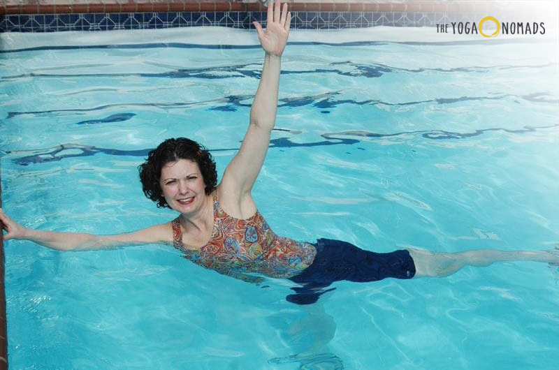 An individual performing a yoga pose in a swimming pool. The person is extending one arm upwards, holding onto a bar or ledge, while the other arm is stretched out to the side. The individual’s legs are extended and slightly separated, with one leg raised higher than the other, creating a dynamic and balanced posture in the water. The water has ripples, indicating movement.