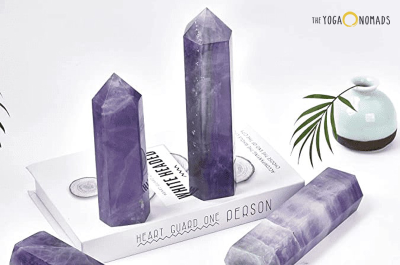 A collection of purple fluorite crystal towers arranged on and around a white book with the title ‘HEART GUARD ONE PERSON’ visible on its spine. The crystals are translucent with varying shades of purple and some white inclusions. The background includes a soft, white surface with subtle shadows, a small green plant in a pot, and an object that appears to be a diffuser or small vase.