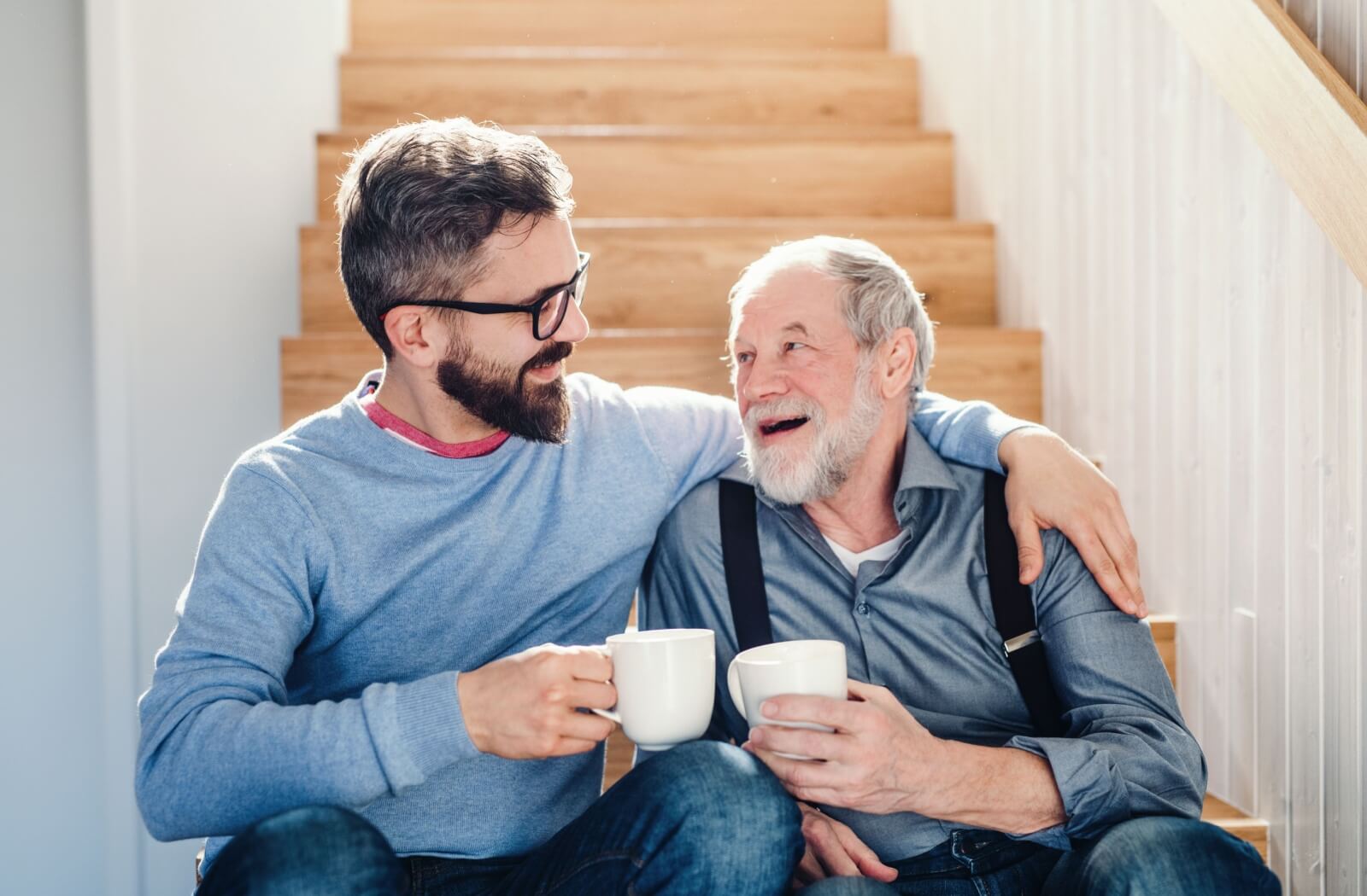 An individual wraps their arm around their older parent while they sit on the staircase with cups of coffee