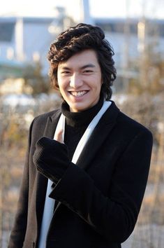 This contains an image of Lee Min Ho wearing a black jacket and white scarf smiles at the camera while holding his hand up to his ear