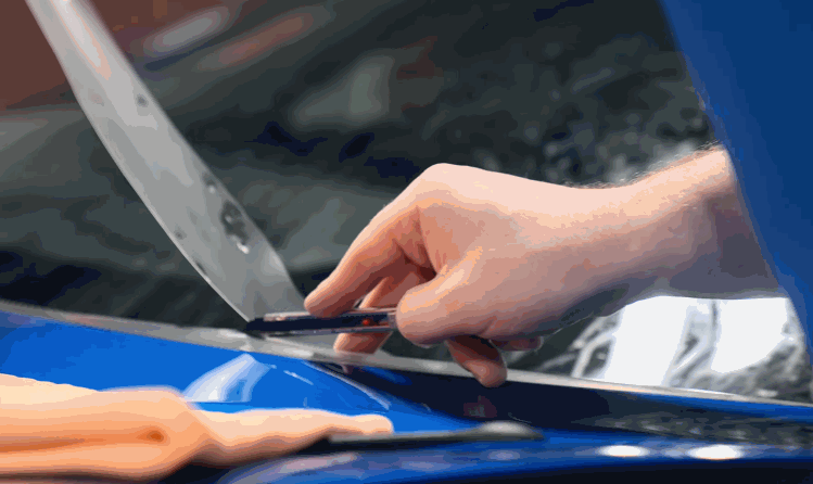 A close-up of a hand using a precision cutting tool to trim a protective film on the edge of a shiny blue car hood, with a microfiber cloth in the foreground