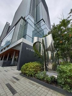 This contains an image of the entrance to hybe building surrounded by greenery and tall buildings in the background