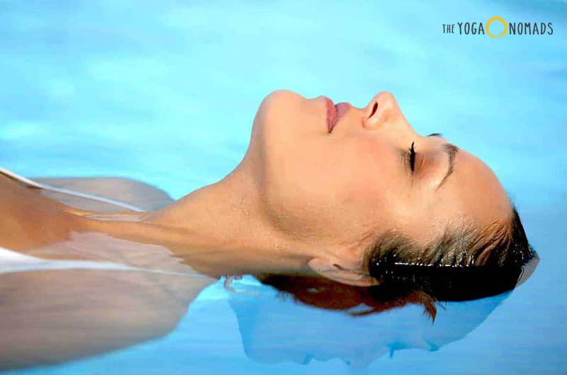 A person’s head partially submerged in clear blue water, creating a reflection on the water’s surface. The individual appears to be practicing a yoga pose or engaging in a relaxation activity in the water. 