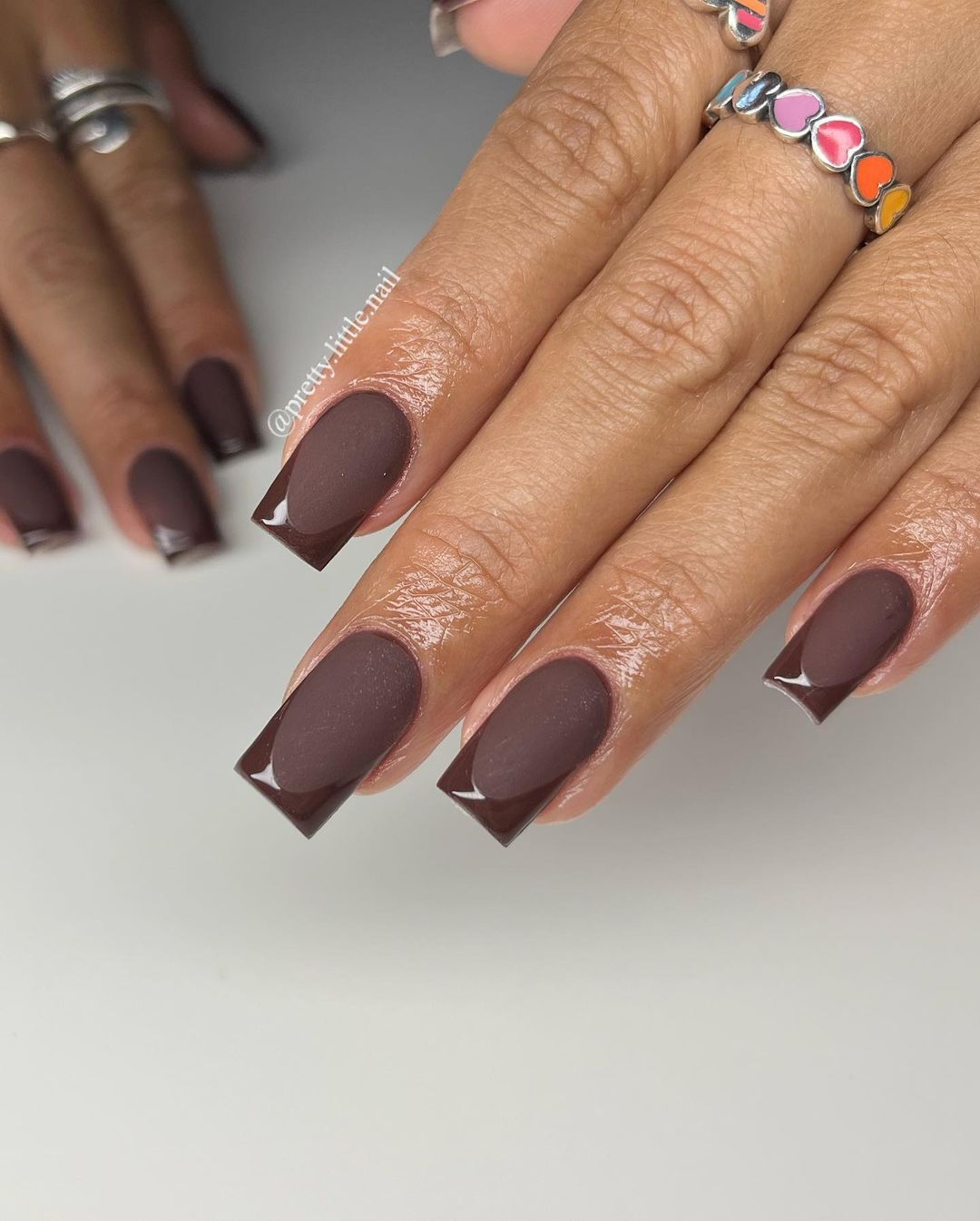 Matte brown nails with glossy tips, featuring heart-shaped nail art on one finger, close-up view.

