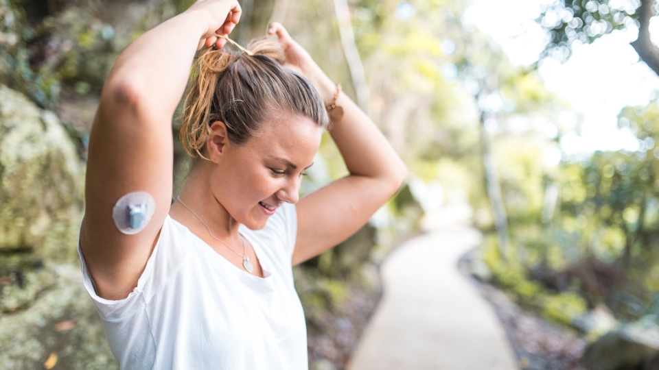 A smiling young woman tying her hair into a ponytail during an outdoor walk, wearing a glucose monitor on the upper part of her left arm.