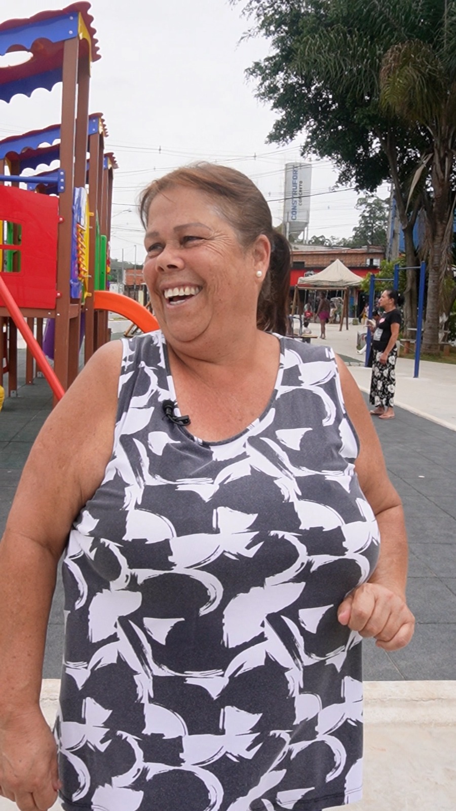 Uma munícipe aparece sorrindo em plano principal da foto, ela veste uma blusa preta e branca. Ao fundo é possível ver grandes brinquedos infantis. 