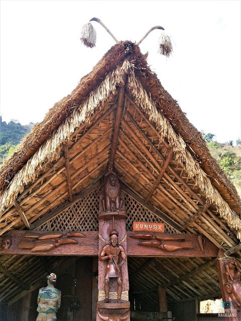 A traditional wooden structure with a thatched roof, featuring intricate carvings of human and animal figures, embodies the essence of Naga houses. Its roof ends are decorated with two protruding elements, reflecting Nagaland architecture.