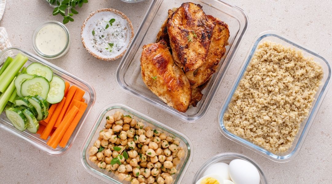 Repas préparés à l'avance avec poulet grillé, quinoa, pois chiches, légumes frais, œufs durs et sauces dans des contenants en verre.