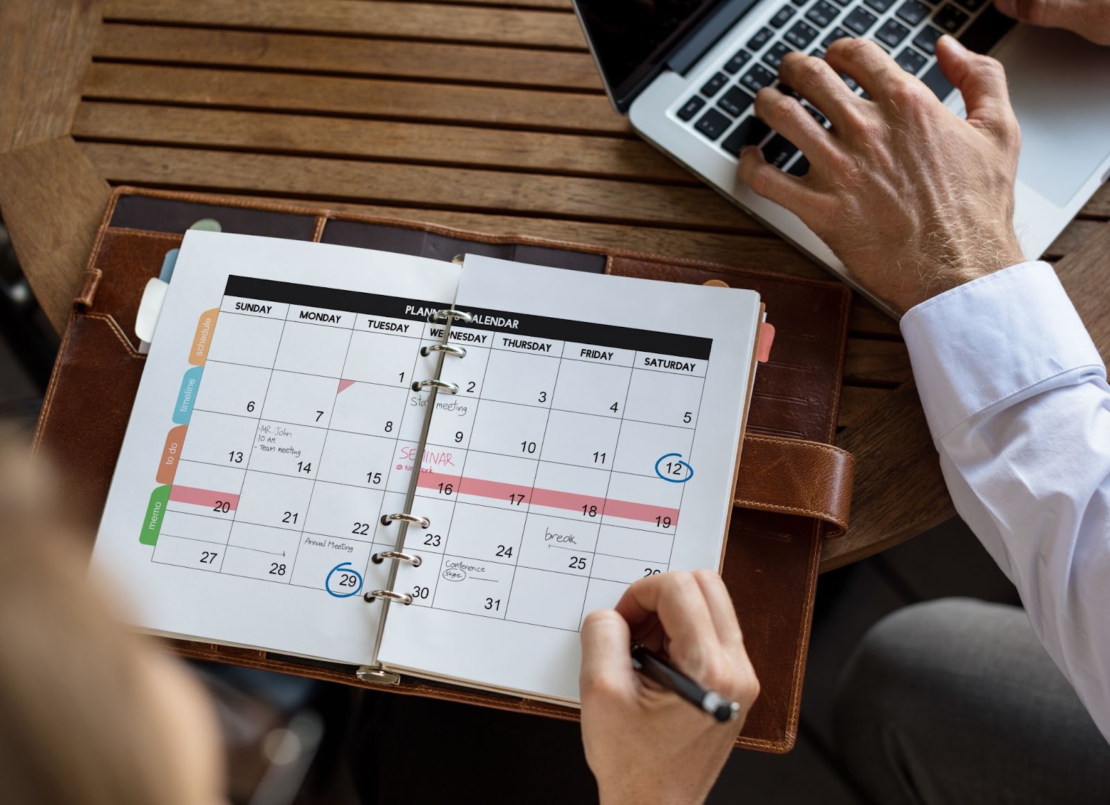 A close-up of the two colleagues’ hands organizing a management schedule; one works on a laptop while the other jots in her planner diary.