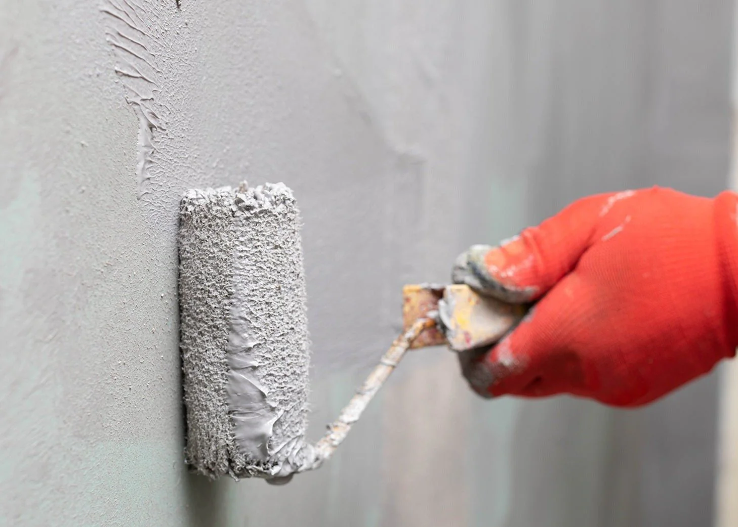 A close-up of a red-gloved hand spreading gray Acriflex pH4 onto a wall with a paint roller