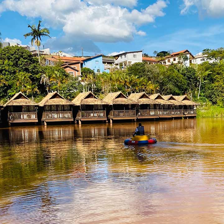 Imagem de uma bela paisagem com casas de palha à beira de um lago, onde uma pessoa navega em uma balsa colorida. Um ambiente tranquilo e natural perfeito para relaxamento.