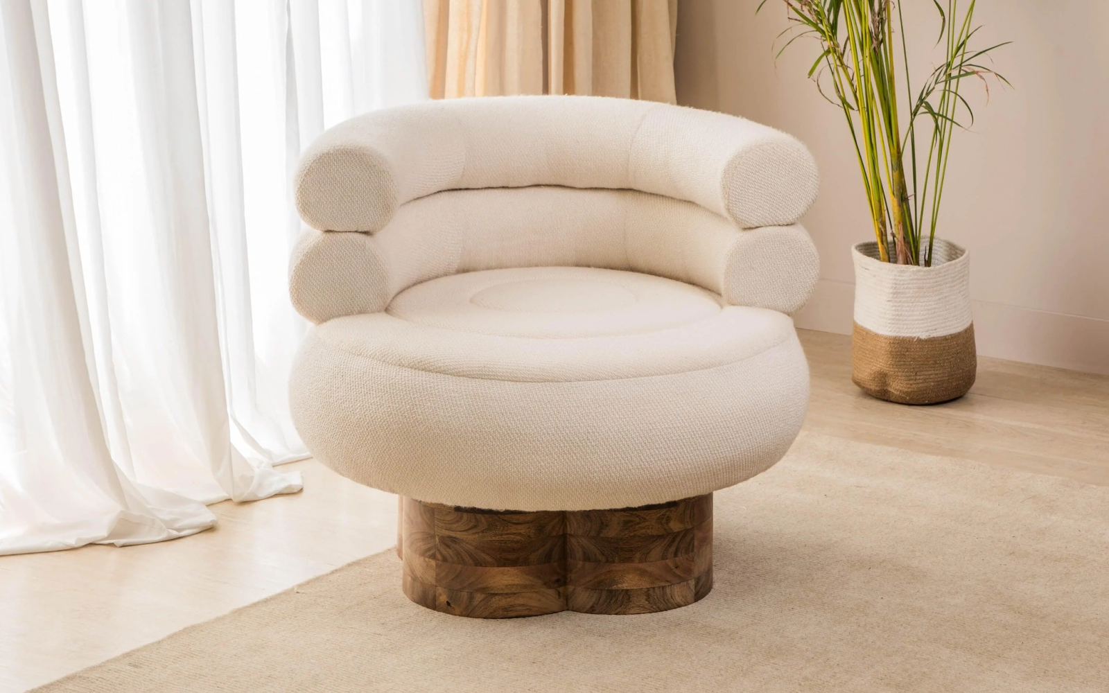 Modern round cream-colored lounge chair with a wooden base and plush cylindrical backrest cushions, placed on a light beige rug near a window with white curtains and a potted plant in the background.