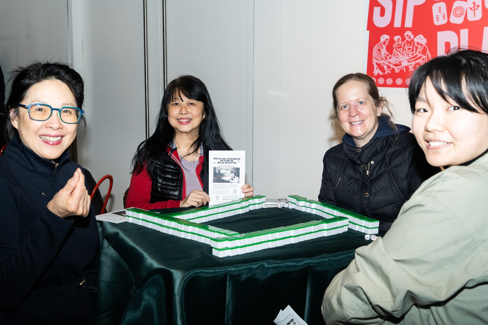 4 women sitting at a mahjong table look into the camera before the game starts. 