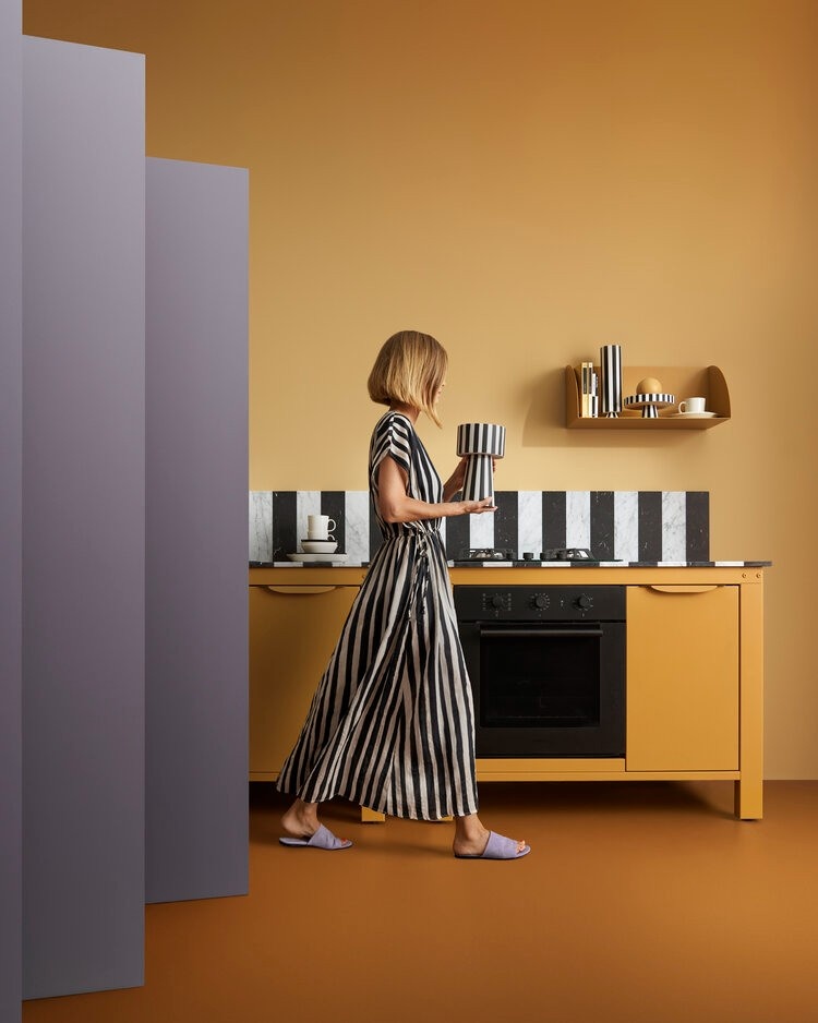 Photography showing a woman in a long, black-and-white striped dress and purple slippers walks in a modern kitchen with yellow cabinets, a black-and-white striped backsplash, and pink walls.