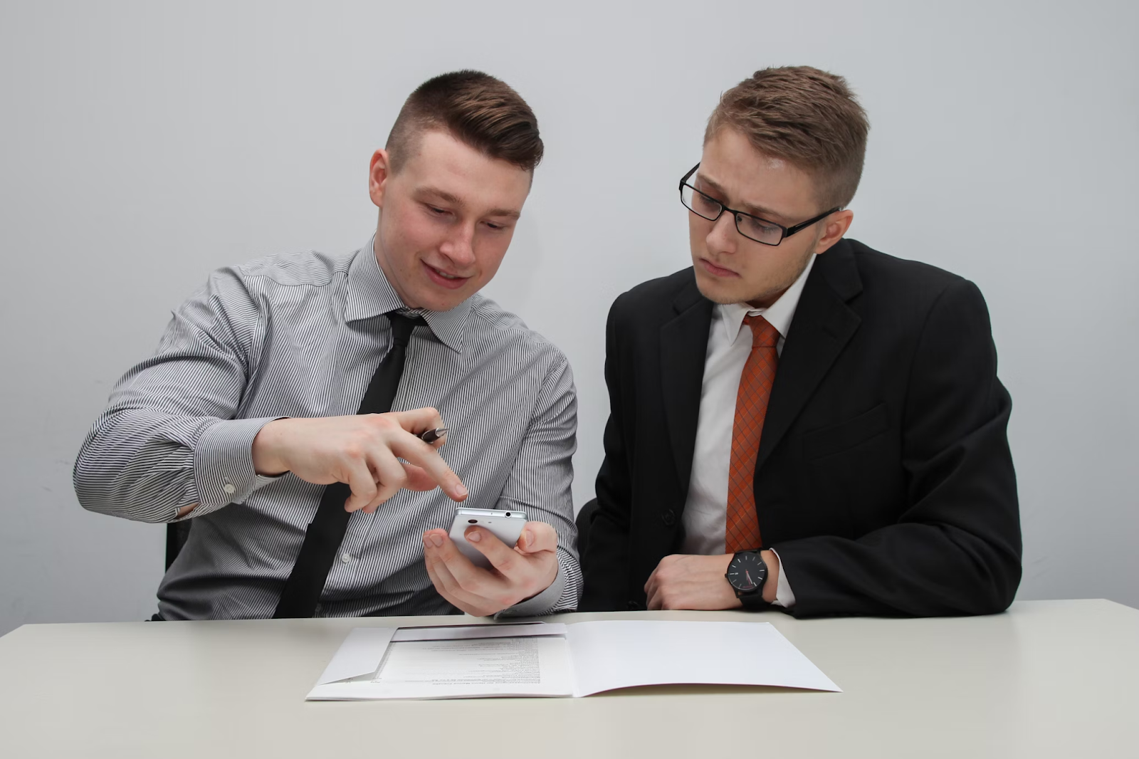 Two businessmen in an office; one discusses resume designations while the other looks at his phone