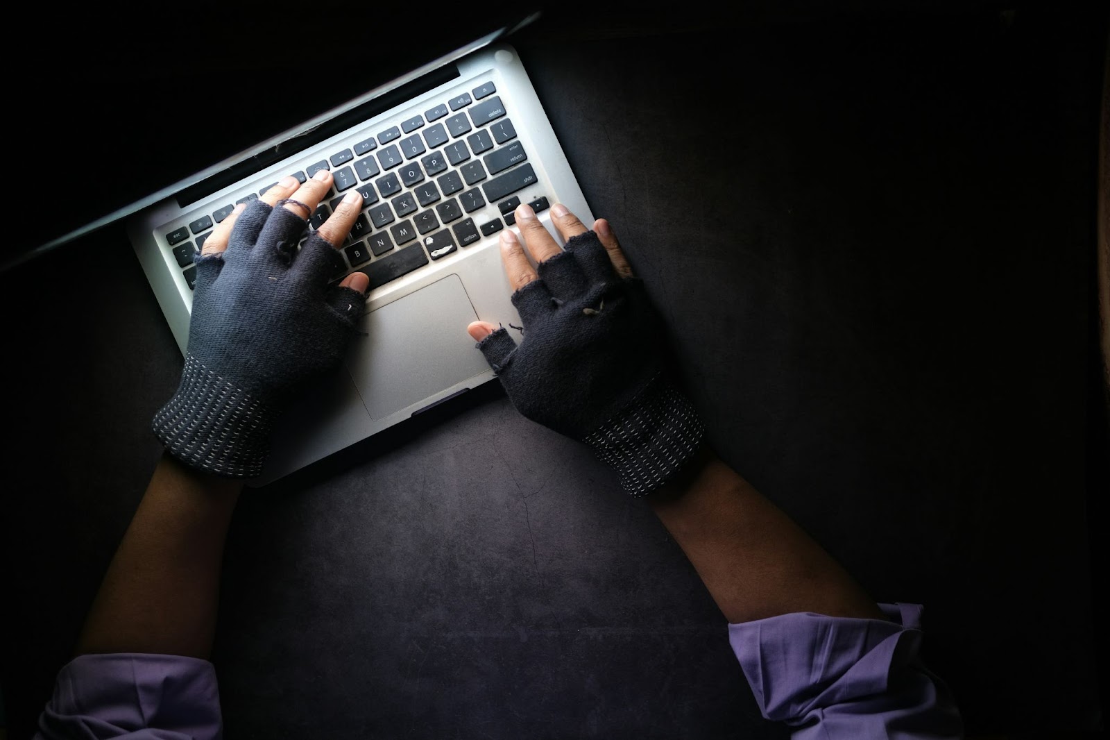 Person wearing fingerless gloves typing on a laptop, representing online scamming activities in a dark setting