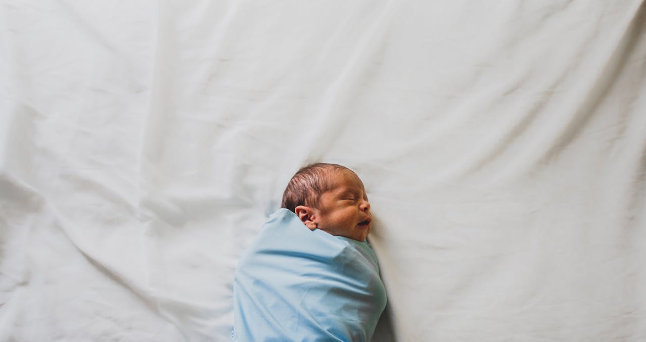 Baby swaddled in blue blanket is sleeping on bed // Healthier Pets Today