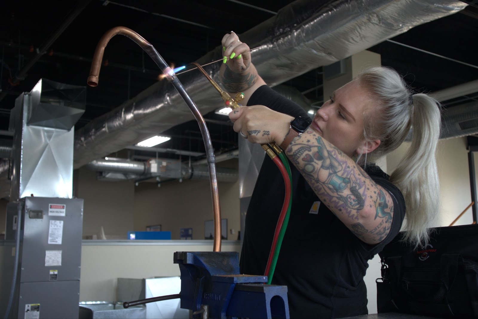 A close-up image of a student working on a copper pipe in an HVAC training setting. The student, with a focused expression, is using a torch to heat the pipe, demonstrating brazing or welding skills. She has a variety of tattoos on her arms and is wearing a black shirt. The background shows industrial HVAC equipment, including ducts and other training setups. The setting captures the hands-on learning environment in the HVAC program.