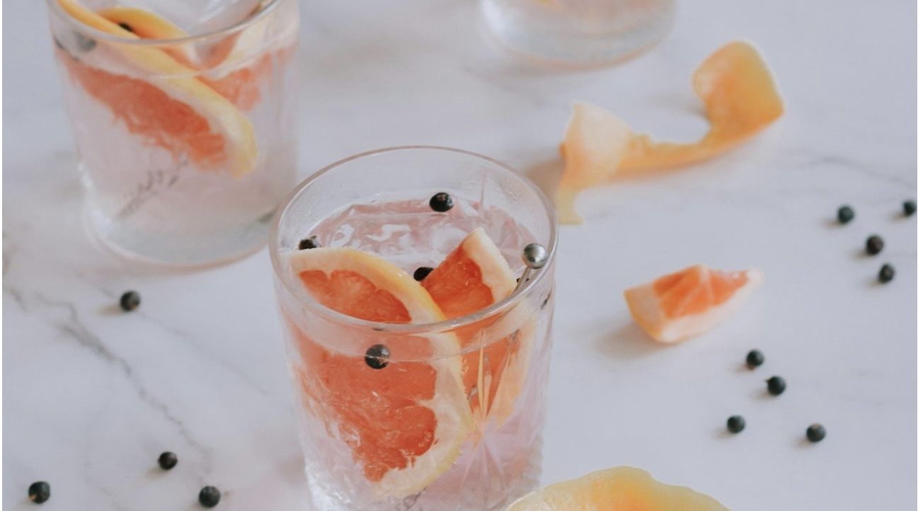 Two gin and tonics are in crystal glasses with grapefruit slices with black peppercorns, and grapefruit peels in the background