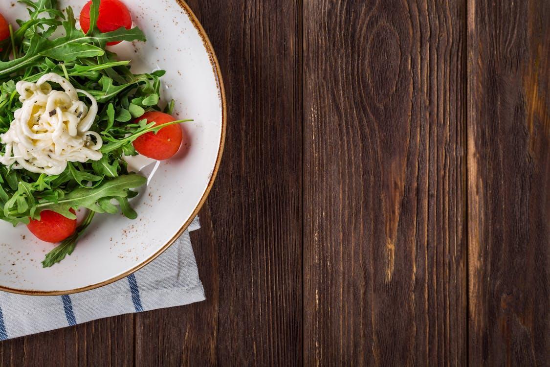 Free A delightful arugula salad with tomatoes and cheese, served on a rustic wooden table, perfect for healthy eating. Stock Photo