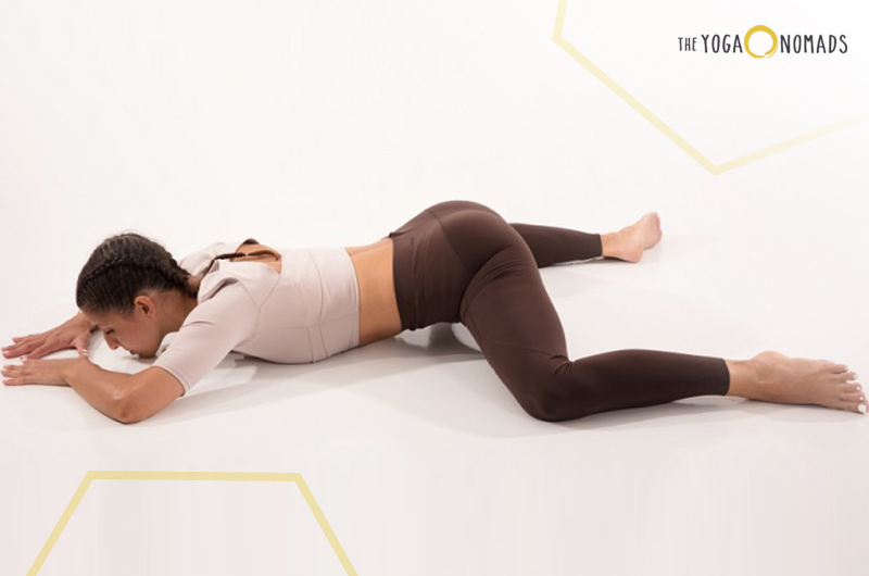 An individual is performing a yoga pose on a mat. The pose appears to be a seated forward fold, with legs spread wide and the torso folded forward. The hands are clasped together. The background transitions from white to pale yellow.