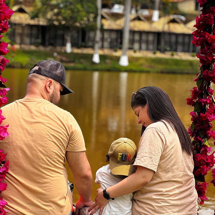 Família ao ar livre, composta por um homem, uma mulher e uma criança, apreciando a natureza perto de um rio, com árvores e uma estrutura de madeira ao fundo.