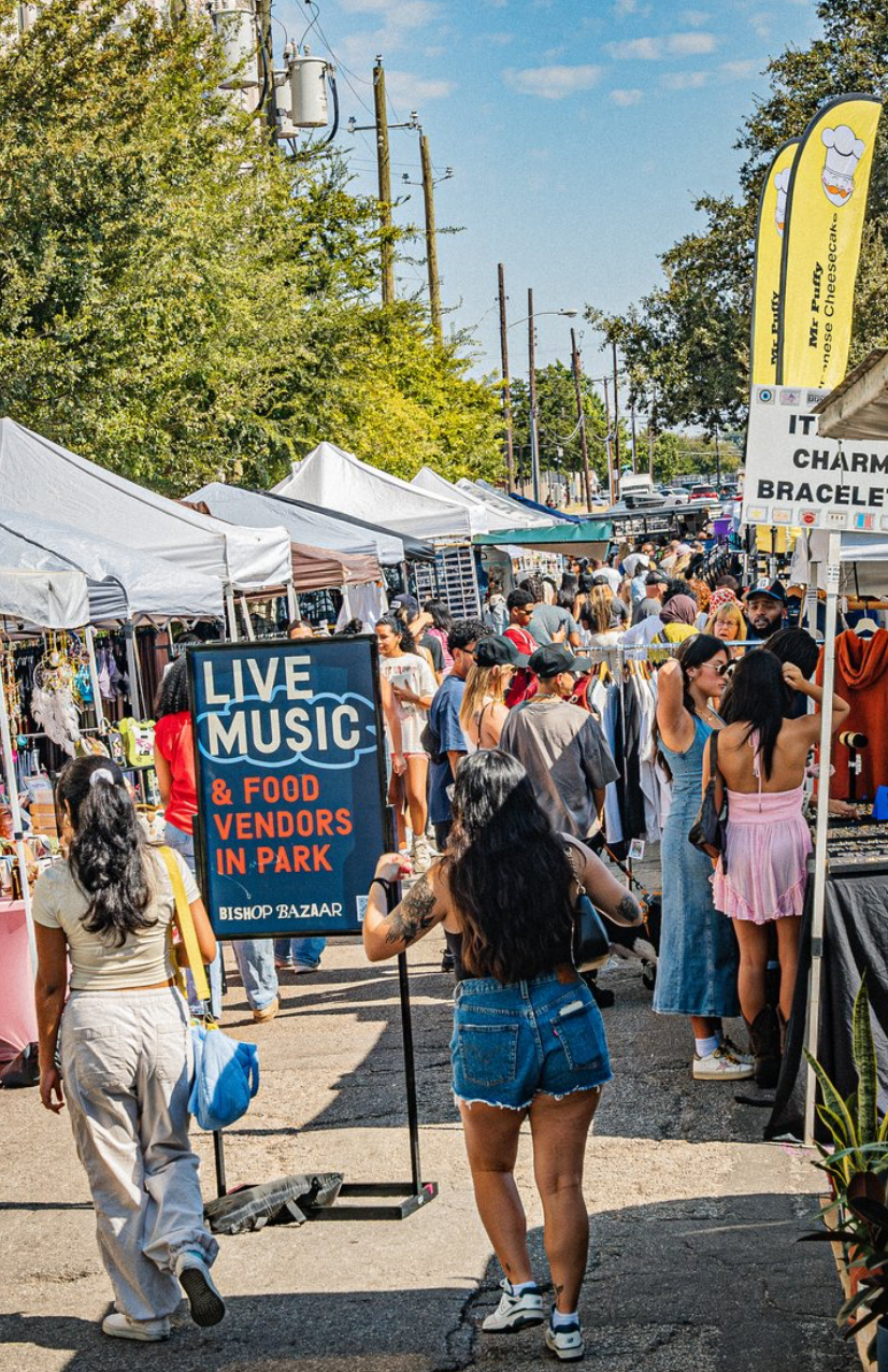 A busy street with vendor tents setup on either side. A sign for Live Music: & Food Vendors in Park is in the center. A large crowd is walking around.