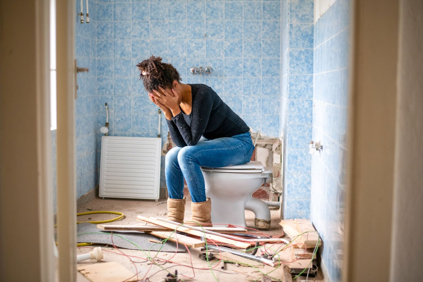 An exhausted homeowner sits on a toilet in an unfinished bathroom, surrounded by water damage and debris from a plumbing emergency caused by broken pipes.