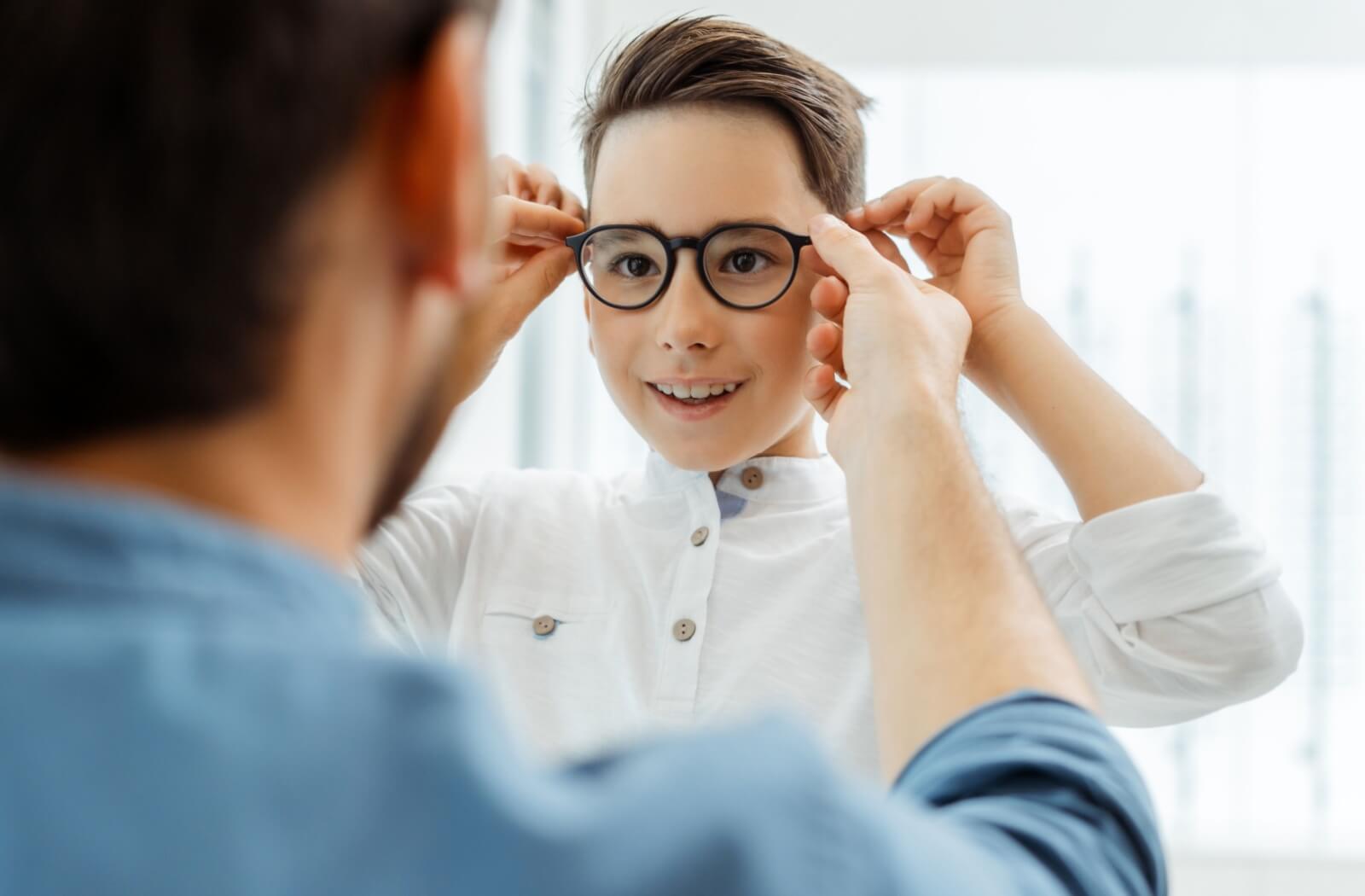 A school-aged child smiling as their parent helps them put on new eyeglasses.