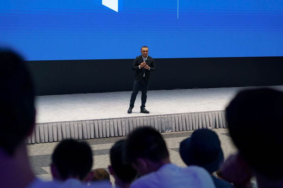 A person on a stage with a large screen behind himAI-generated content may be incorrect.