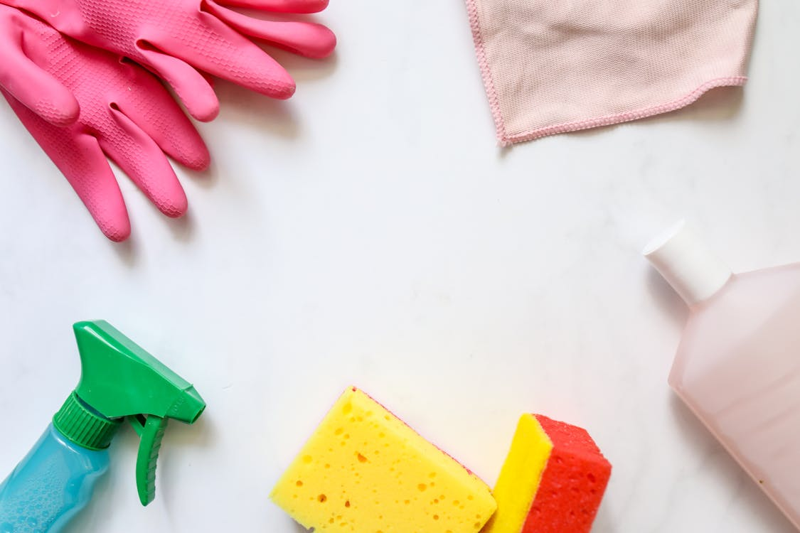 Cleaning supplies arranged on a white surface, including pink rubber gloves, a green spray bottle, yellow and red sponges, a pink cleaning cloth, and a bottle of cleaning solution, emphasizing household cleaning tools