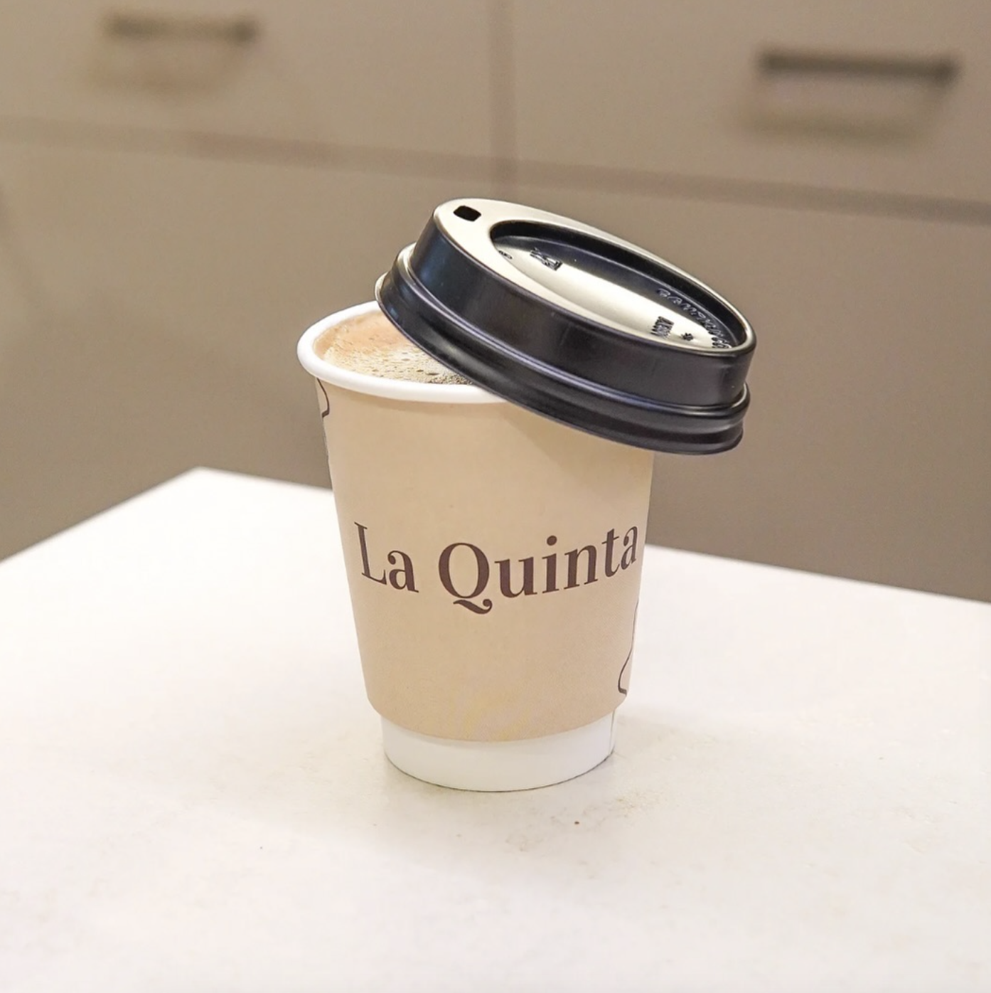 Branded paper coffee cup with black lid, featuring 'La Quinta' logo, containing a hot beverage, placed on a light-colored surface.