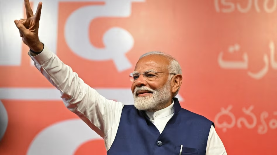 Prime Minister Modi smiling and waving to the crowd at a rally, greeting them with a warm gesture.