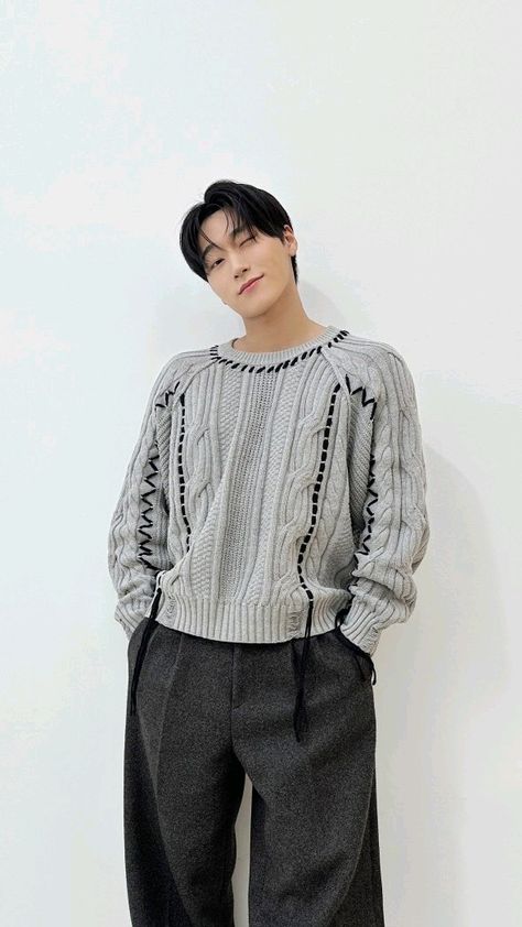 This contains an image of ATEEZ's San  standing in front of a white wall wearing black pants and a sweater