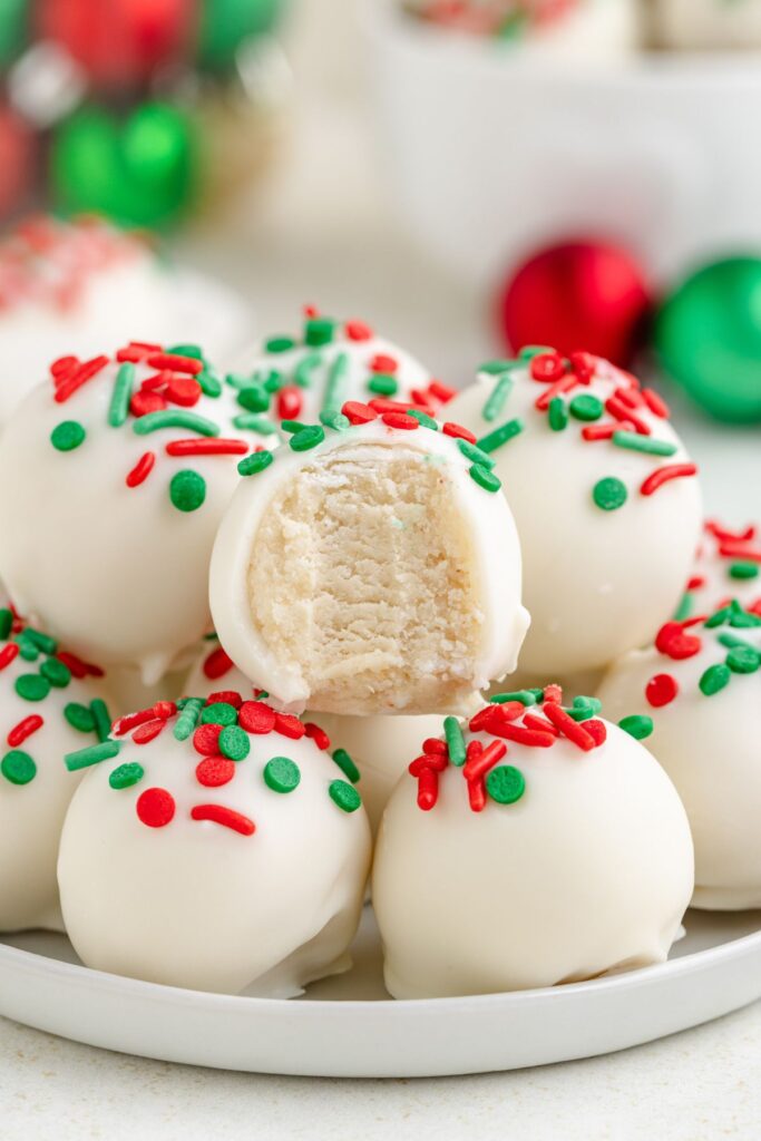 A cock with white truffles topped with red and green sprinkles. One trumel is partially eaten by showing a creamy interior. Christmas decorations are blurred in the background.